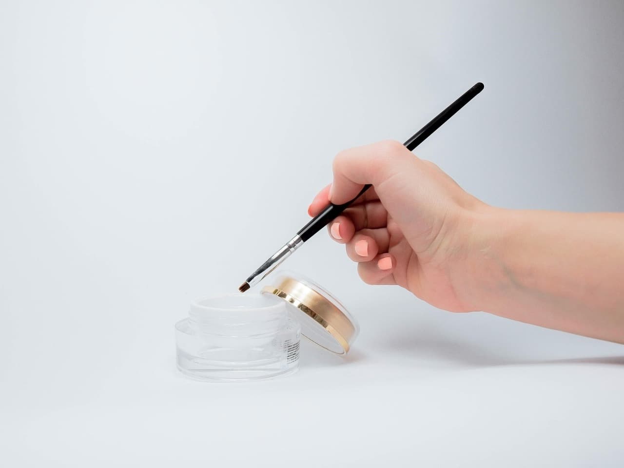 A hand with peach nails dipping a fine nail art brush into a clear gel pot