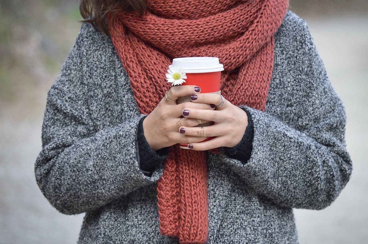 Hands with dark berry nails and rings holding a takeaway cup, wrapped in a knitted scarf