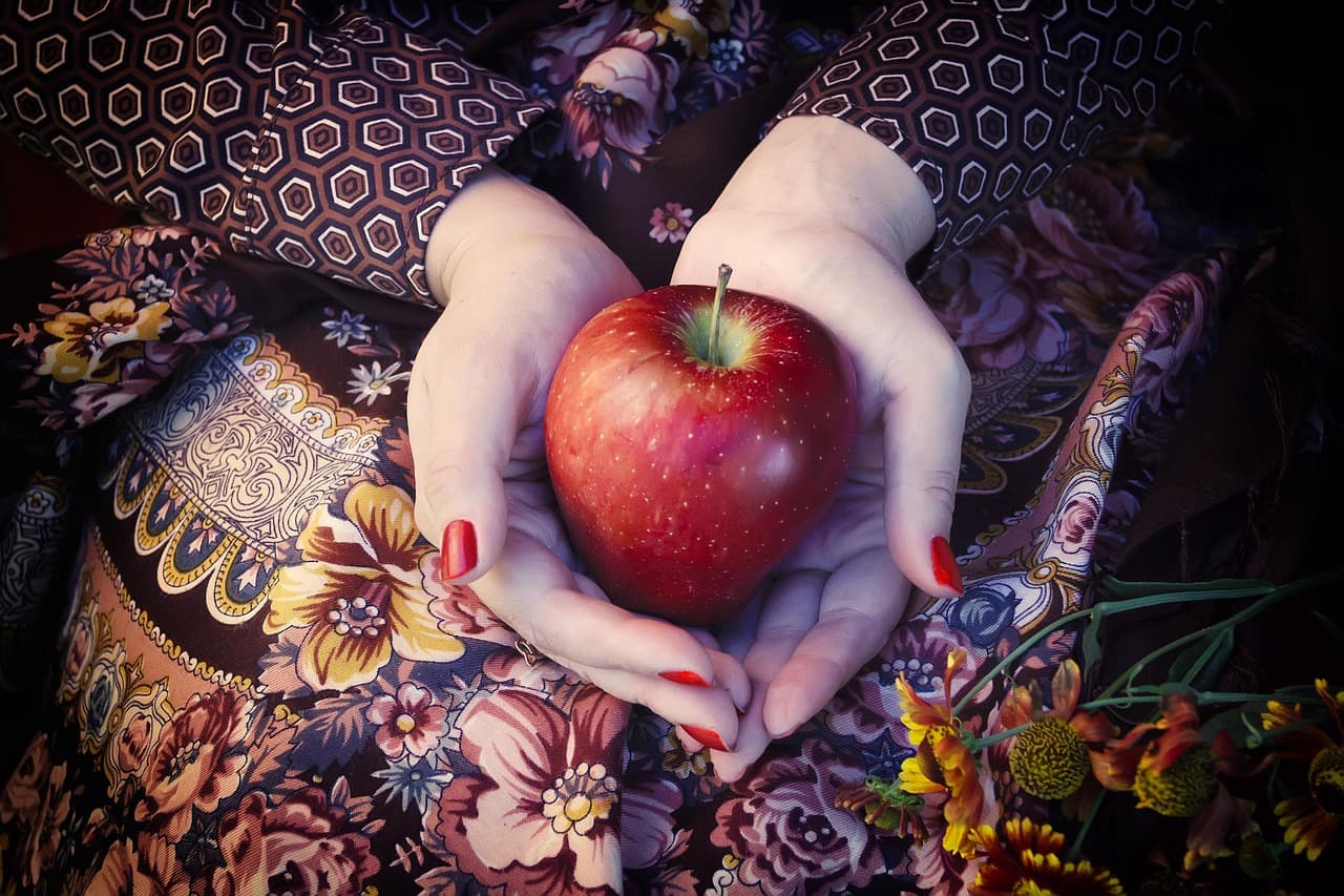 Hands with glossy red nails holding a red apple against a floral dress
