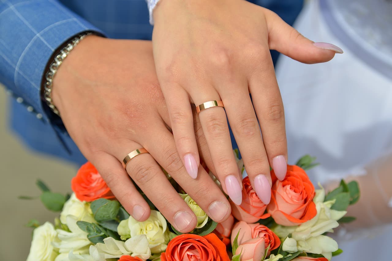 A bride and groom's hands with wedding rings and long almond nails in a soft pink, over a bouquet of roses